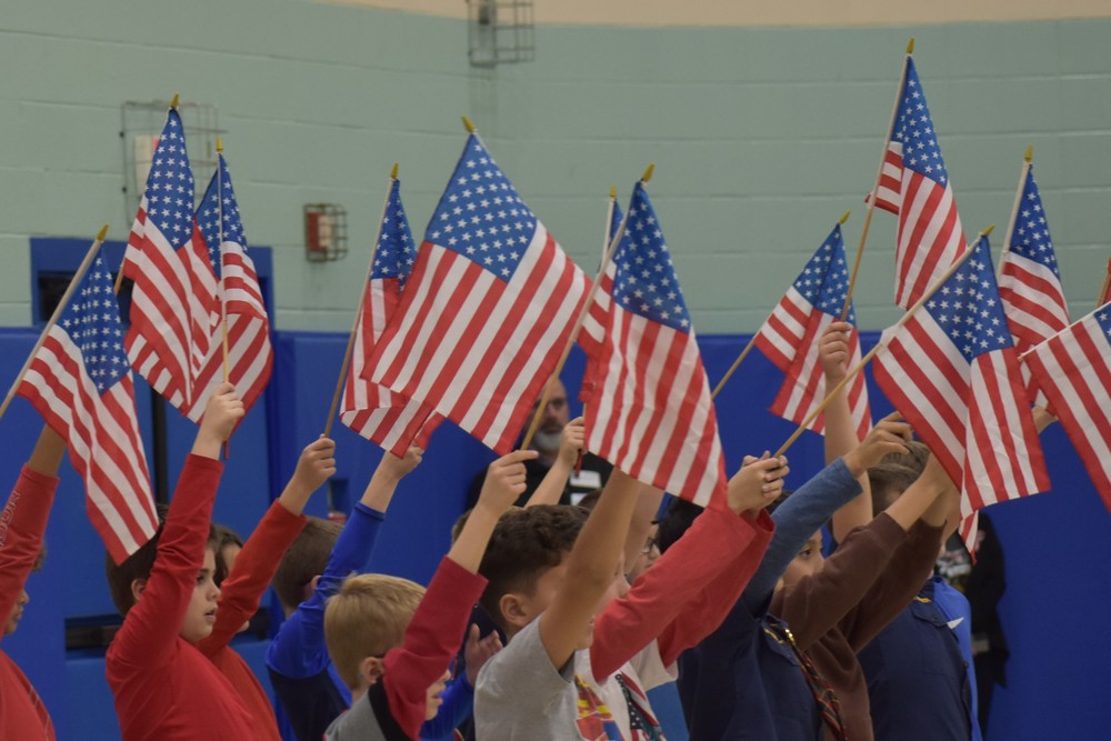 Elementary students hold American flags up above their heads while singing a patriotic song in the Evergreen large gym
