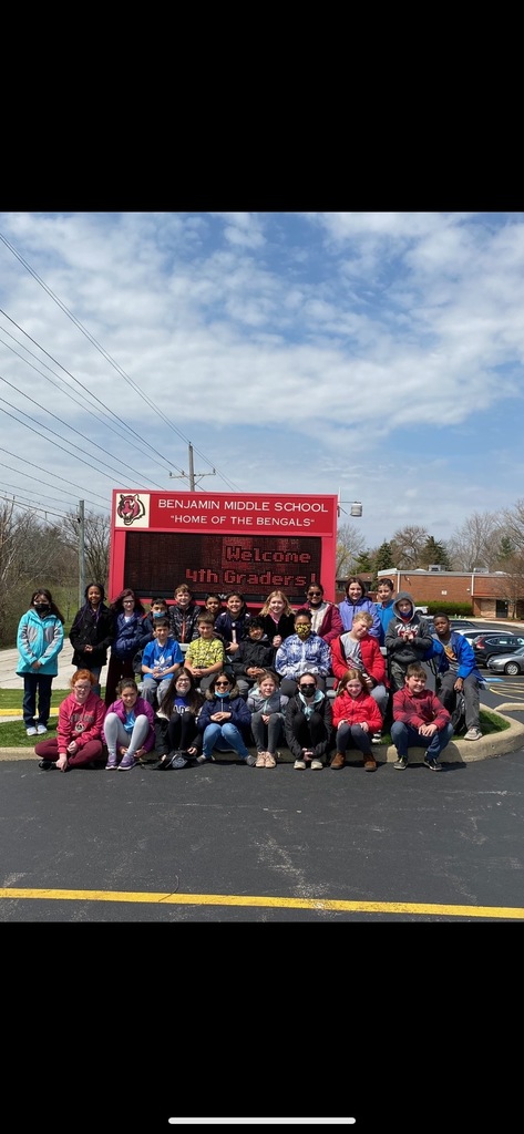 4th grade students in front of the Benjamin Middle sign. Three rows of students. First row is sitting on the ground,  second row is  sitting on bench, third row is standing. Red school sign behind them.  Blue sky with white clouds. 