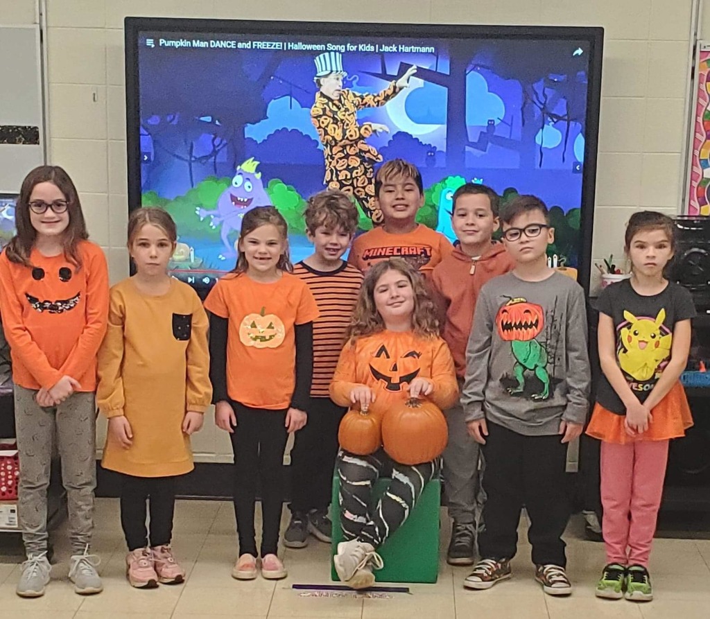 nine elementary students standing in a row, one is sitting, donning pumpkin or fall theme/color shirts 