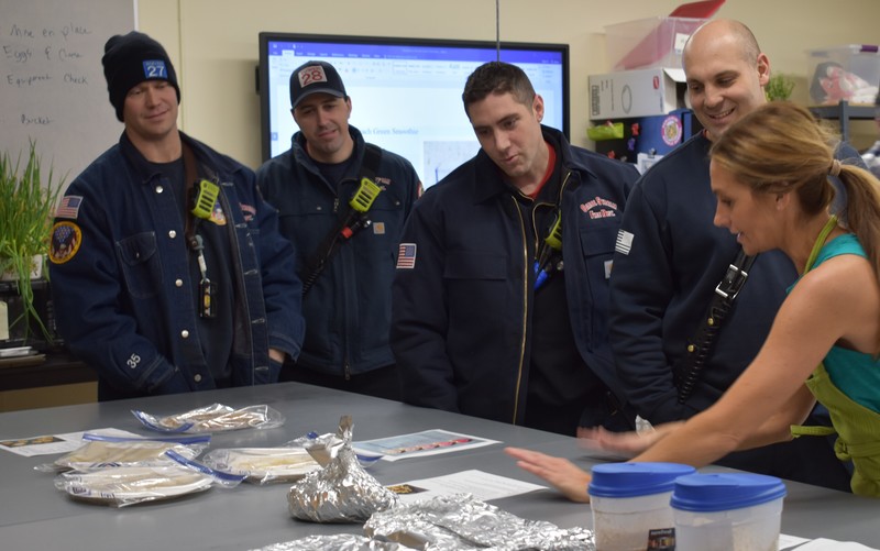 Four firefighters look at packaged food on a table while Mrs. Vidic gestures toward the recipe sitting next to it.