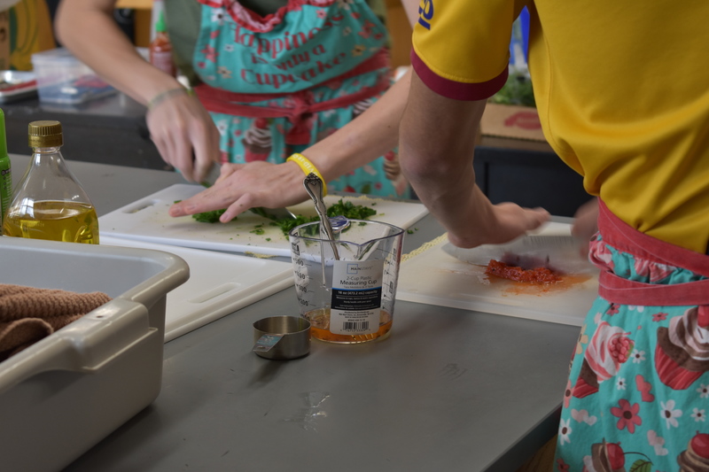 Students hands use large knives to mince vegetables on white cutting boards.