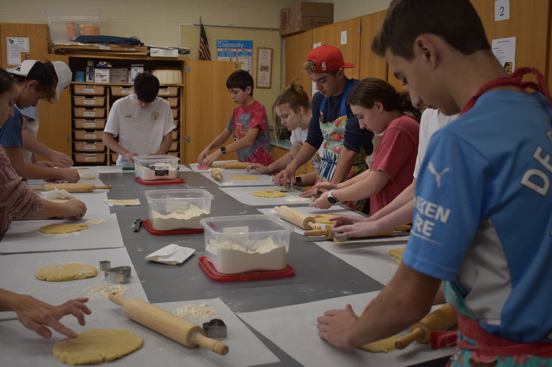 Students stand around a table with rolling pins and discs of cookie dough ready to roll out.