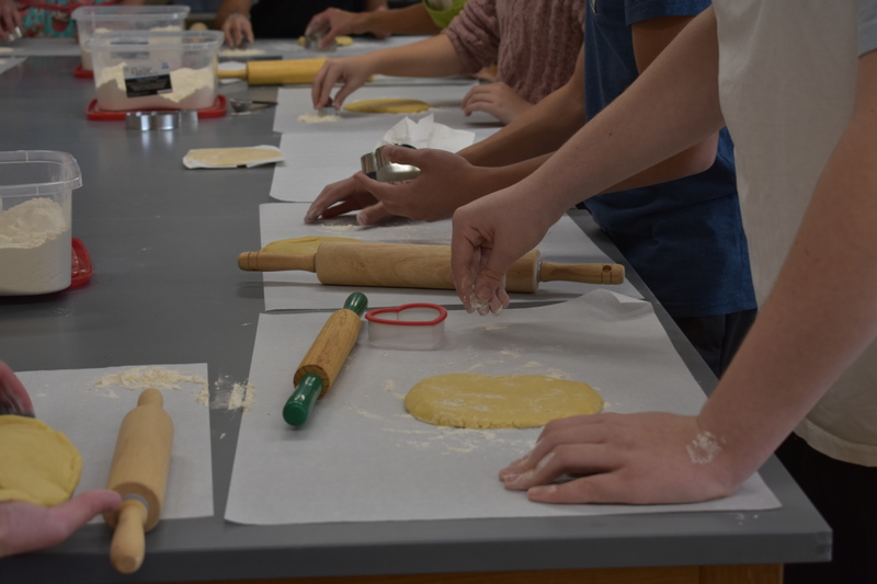 Student hands sprinkle flour over a disc of rolled out cookie dough.