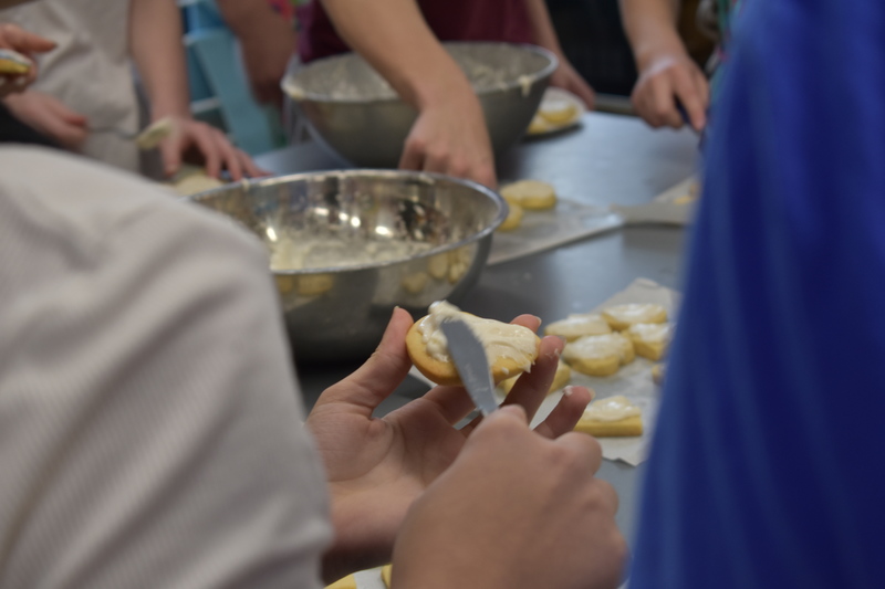 Student hands hold a heart shaped sugar cookie and spread frosting on the top of it with a butter knife.