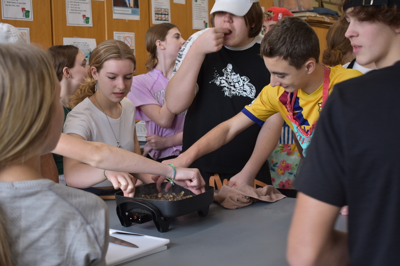 Seven students gather around an electric skillet and grab mushroom pieces to taste.
